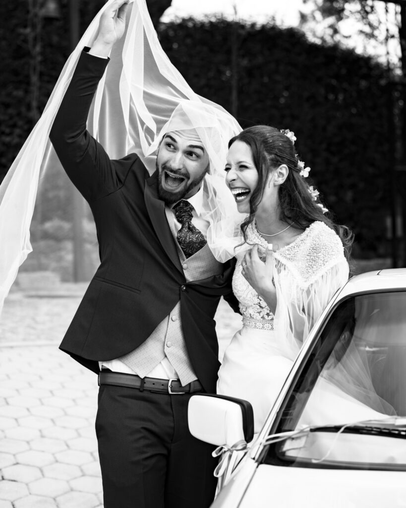 Noivos em gargalhada segurando o véu ao vento junto a carro clássico branco, momento de alegria em preto e branco.