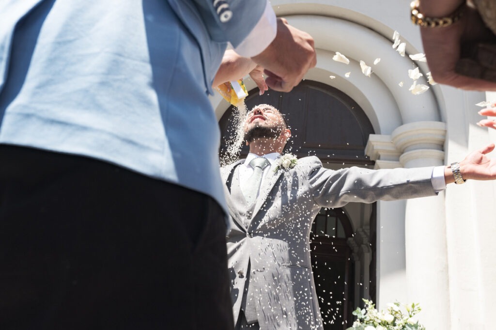Noivo de braços abertos recebe chuva de arroz à saída da igreja, contraluz solar forte, momento de celebração espontâneo.