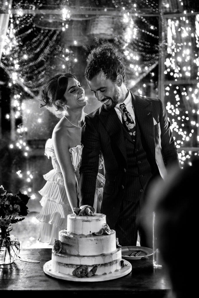 Noivos cortam o bolo alegremente, estando a noiva a sorrir em direção ao noivo, registo documental a preto e branco. 