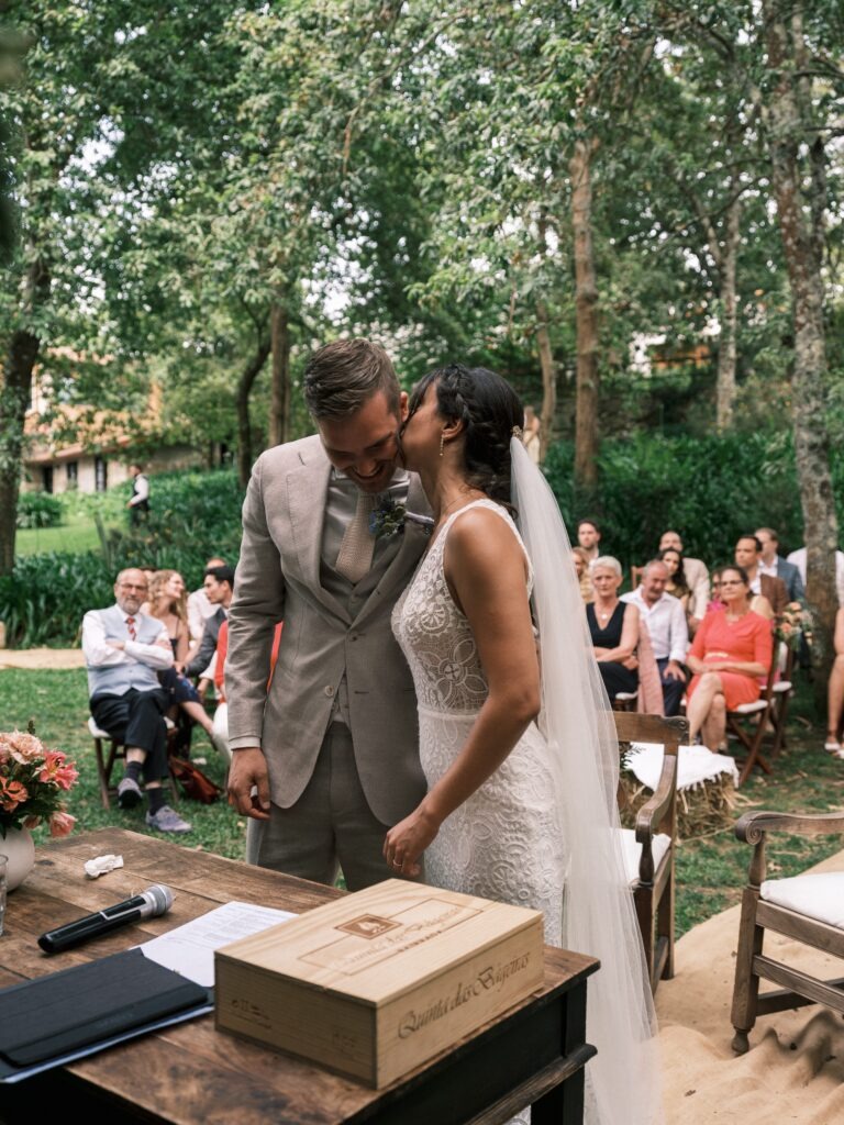 Noiva da beijo na face do noivo, enquanto este se ri e ambos são observados pelos seus convidados durante a cerimónia. 