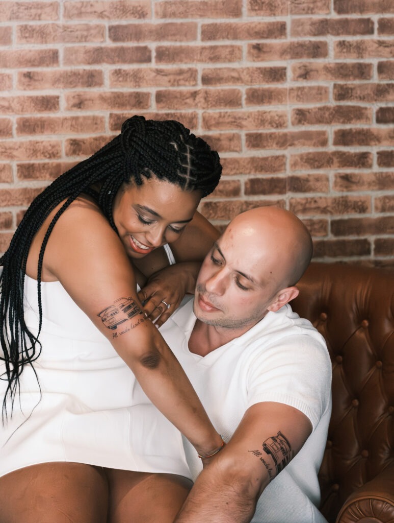 
Casal de noivos a mostrar a tatuagem feita na sessão de solteiro, estando o noivo sentado num cadeirão enquanto a noiva está sentada no colo dele, onde ambos estão a observar a tatuagem dela, sendo igual à dele.