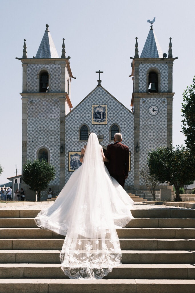 Noiva e pai de costas para o fotógrafo, subindo os últimos degraus das escadas em direção à igreja. 