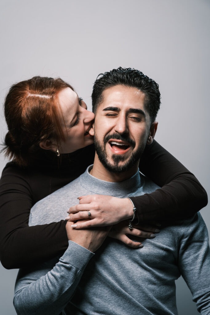 Casal em sessão de pré-casamento dentro do estúdio com fundo branco, a noiva morde a orelha do noivo, enquanto este faz uma careta. © Filipe Santos 2025
