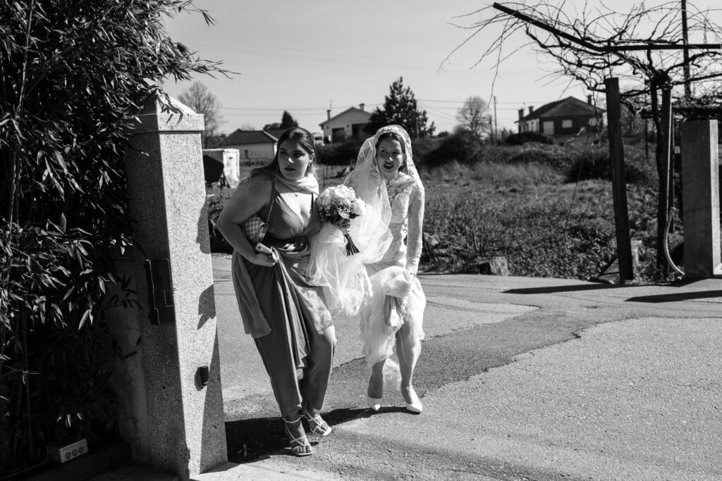 Noiva a caminho da igreja, segurando no fundo do seu vestido, enquanto a madrinha segura-lhe no véu e no ramo das flores, imagem a preto e branco