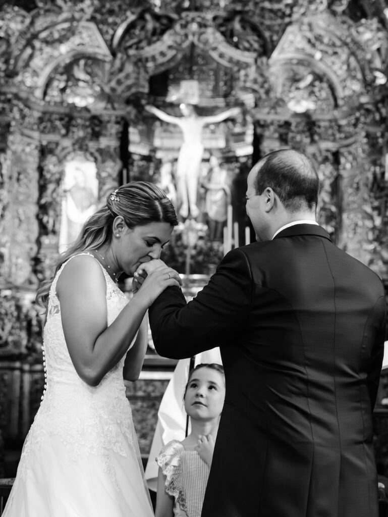 Noiva beija a aliança do noivo durante a cerimónia de casamento, registo documental a preto e branco.