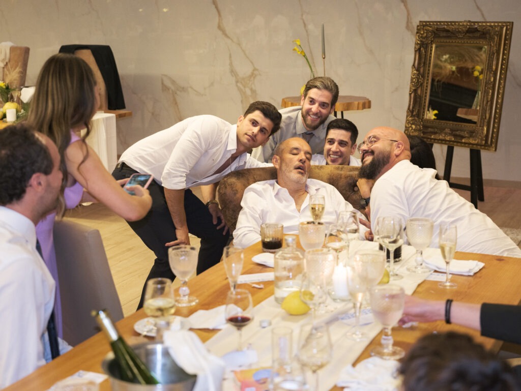 Momentos engraçados entre convidados, estando eles a fotografar outro convidado que adormeceu num cadeirão enquanto outros ficam à volta dele a sorrir. 