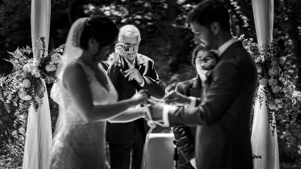 Noivos a atar as mãos na cerimónia de handfasting com convidado a fotografar em câmara analógica ao fundo, altar de flores, preto e branco.