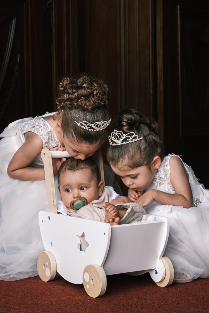 Duas meninas com tiara a espreitar o irmão bebé num carrinho de brincar, momento terno no interior, instantes antes da entrada na igreja em Aveiro, registo documental.