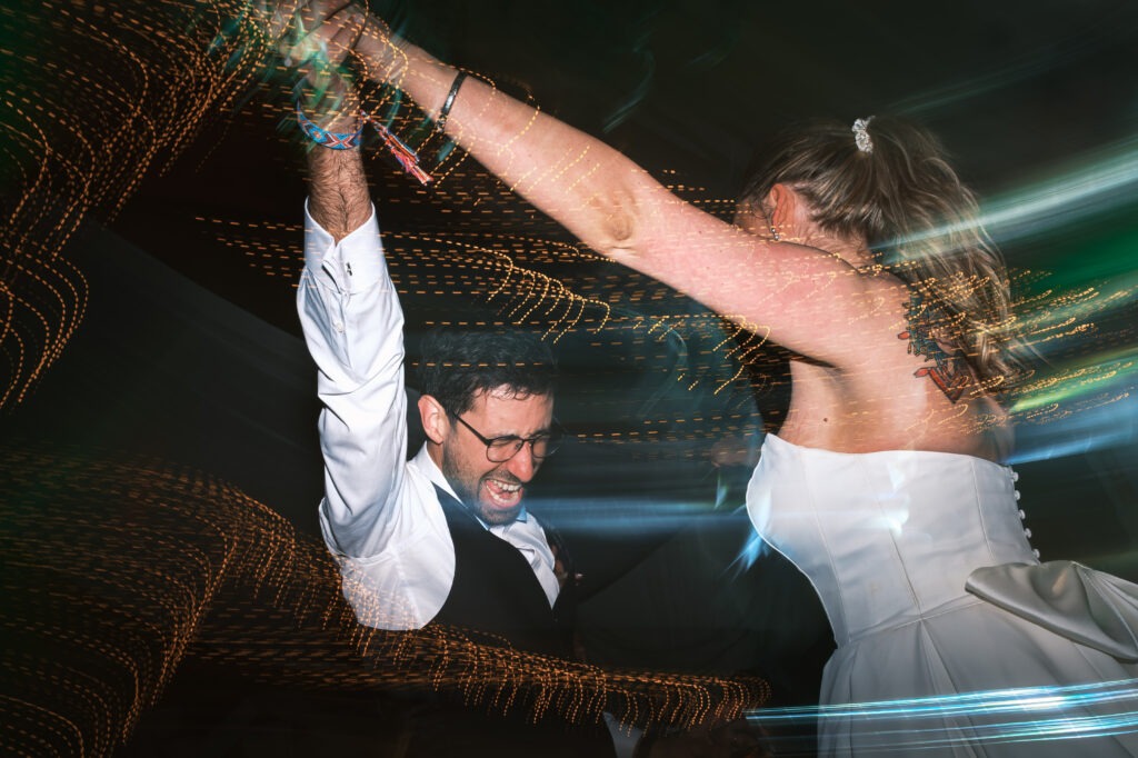 Noiva e noivo a dançar na pista; traços de luz criados com flash e arrasto de obturador, energia e festa com convidados desfocados ao fundo. 