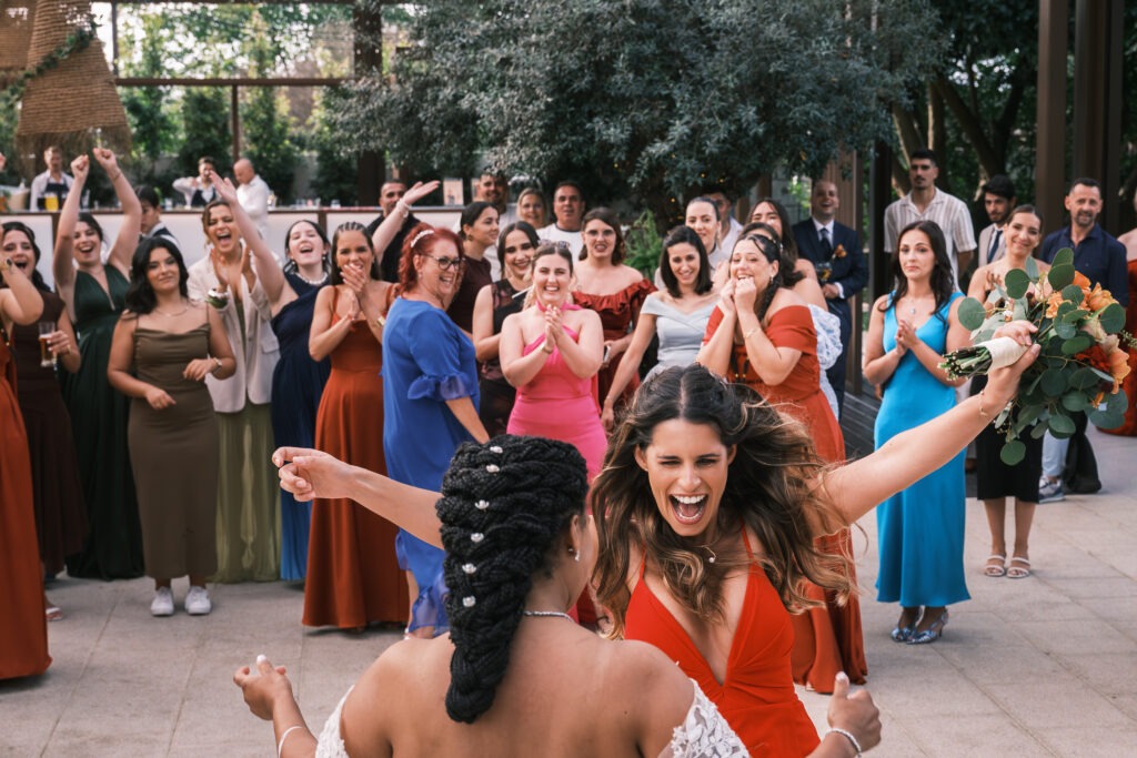 Amiga que apanhou o bouquet corre para a noiva com braços abertos, convidados a vibrar ao fundo, momento espontâneo ao ar livre no Porto, registo documental.