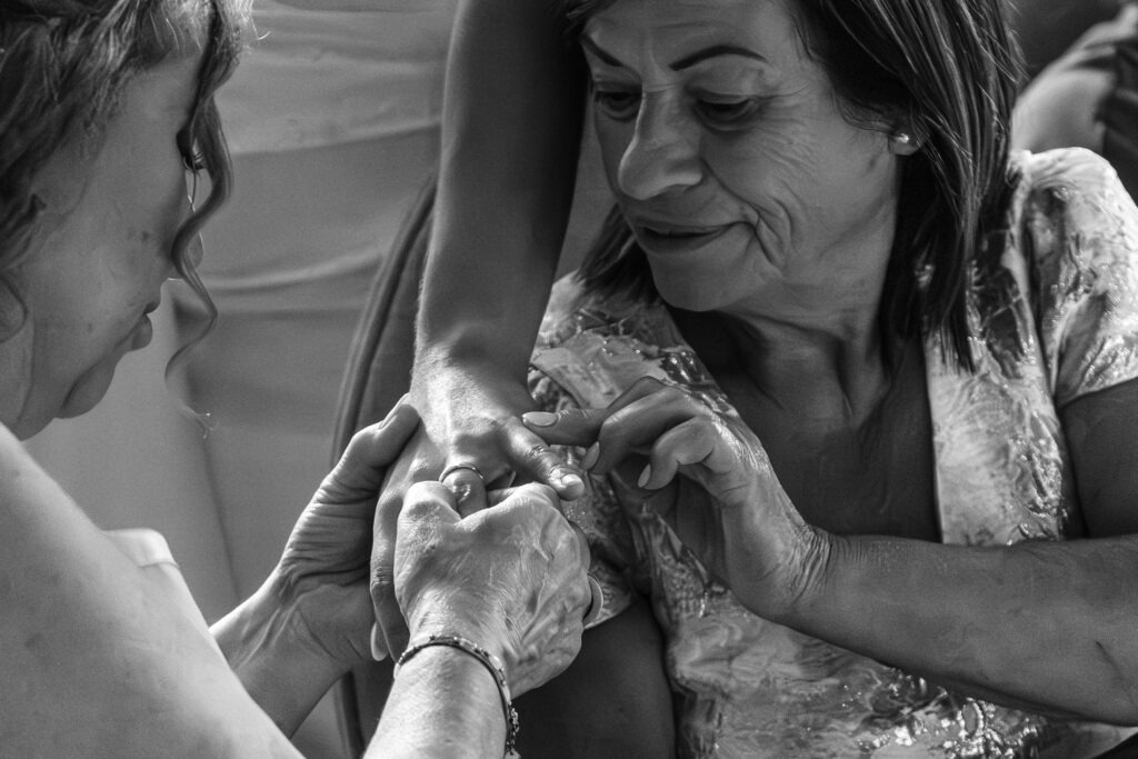 Close-up das mãos enquanto familiares observam a aliança no dedo da noiva; ternura e gesto de carinho em registo documental a preto e branco no Porto.