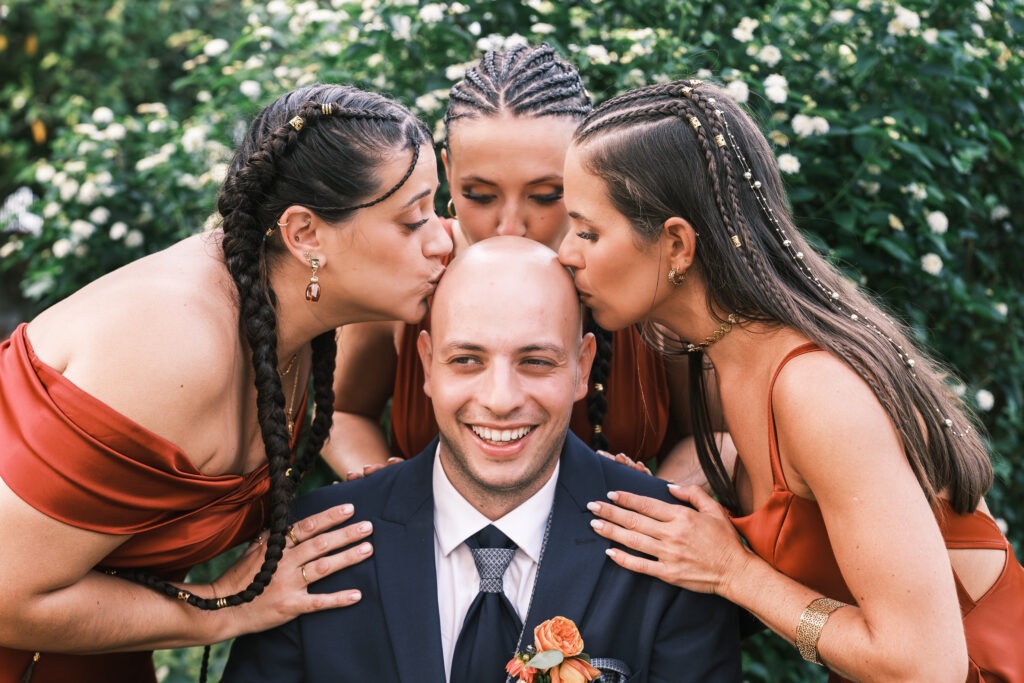 Madrinhas a beijar a cabeça do noivo num momento divertido no jardim, risos e cores terracota, registo documental em Porto.