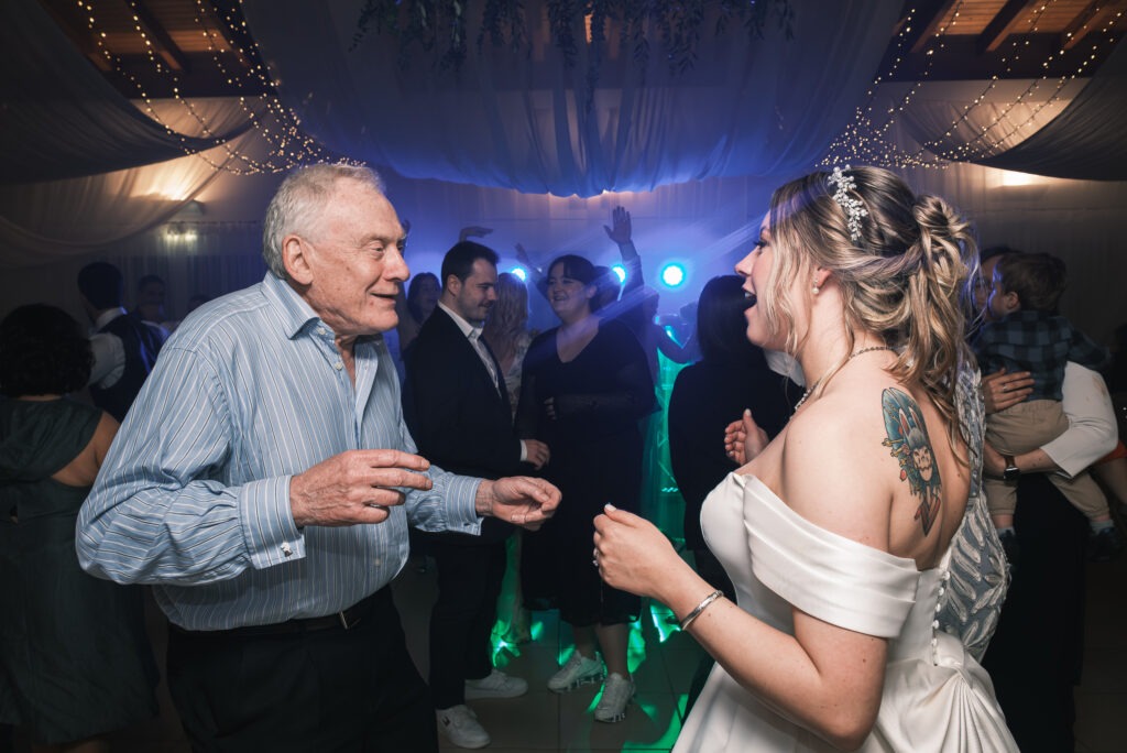 Noiva a dançar com o avô na pista de dança, luzes de DJ e convidados ao fundo; momento terno e divertido, registo documental no Porto.