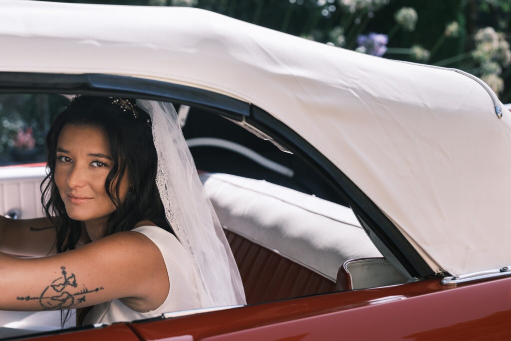 Noiva dentro de carro clássico vermelho, olhar cúmplice para a câmara, véu visível, momento antes da cerimónia no Porto, registo documental.