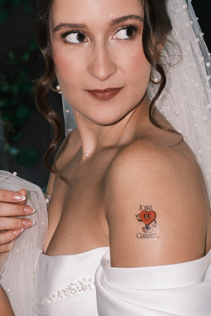 Close-up do ombro da noiva com tatuagem do casamento em forma de coração, véu e detalhes com pérolas, luz de flash suave, registo documental no Porto. 