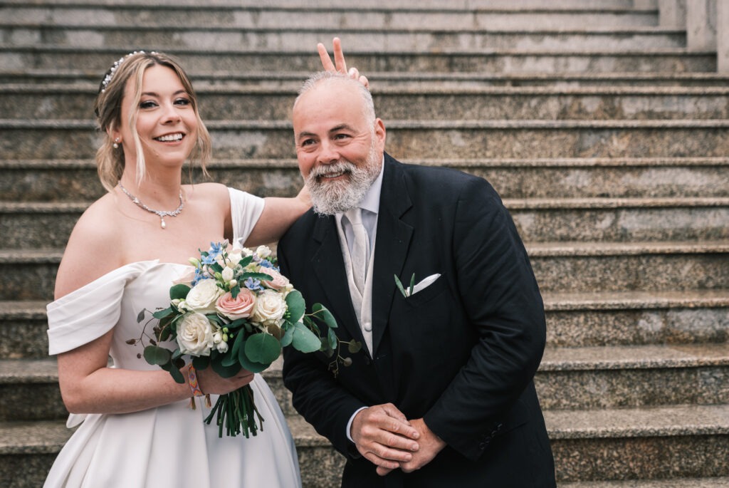 Noiva com bouquet abraçada ao pai nas escadas da igreja; gesto divertido com “orelhas” atrás dele, sorrisos e ambiente de celebração, registo documental no Porto