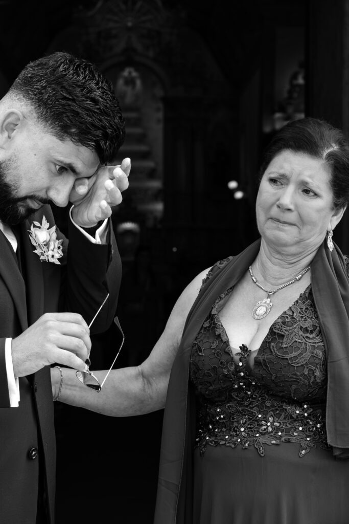 oivo a limpar uma lágrima enquanto segura a mão da mãe à porta da igreja, instante emotivo antes da cerimónia em porto, registo documental a preto e branco.