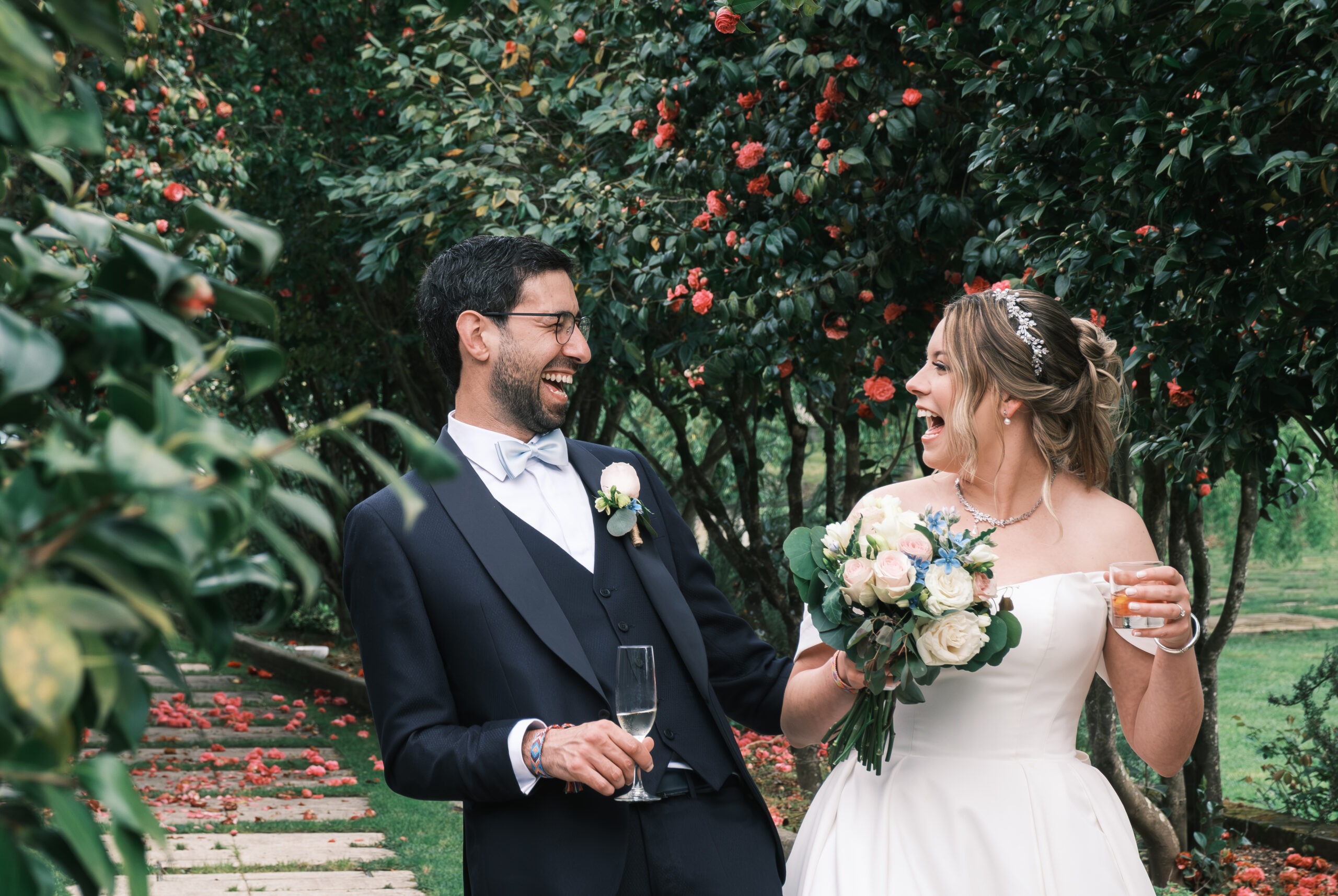 Noivos a rir com bebidas na mão durante os aperitivos na quinta, bouquet em destaque, registo documental ao ar livre