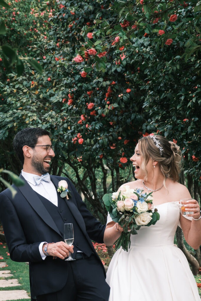 Noivos a rir com bebidas na mão durante os aperitivos na quinta, bouquet em destaque, registo documental ao ar livre