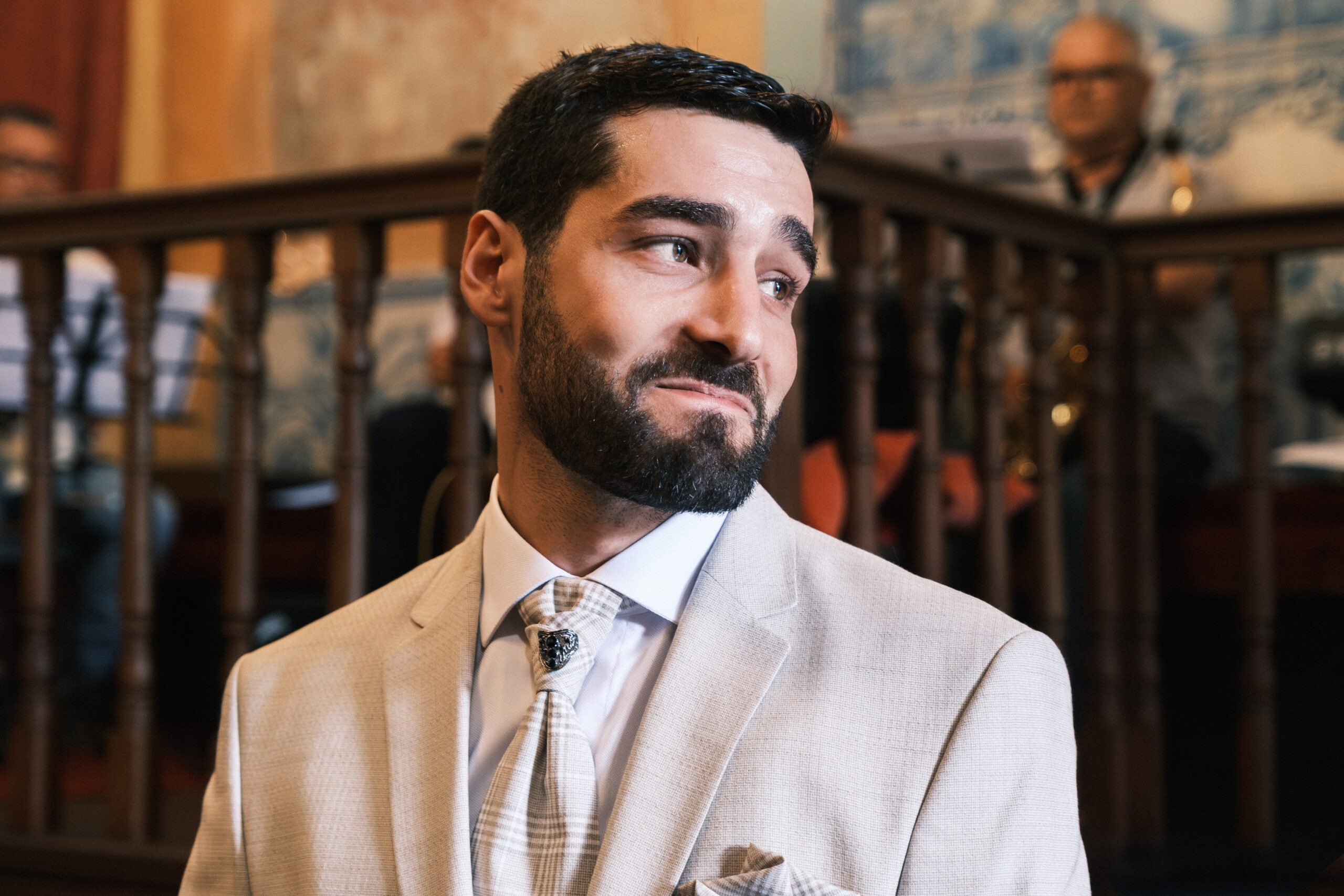 Retrato do noivo durante a cerimónia, olhar de lado com expressão de expectativa; luz natural em interior, Portugal.