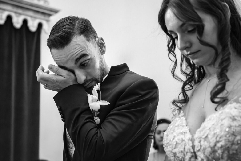 Noivo limpa uma lágrima durante a cerimónia; noiva ao lado, emoção em registo documental, preto e branco, Portugal.
