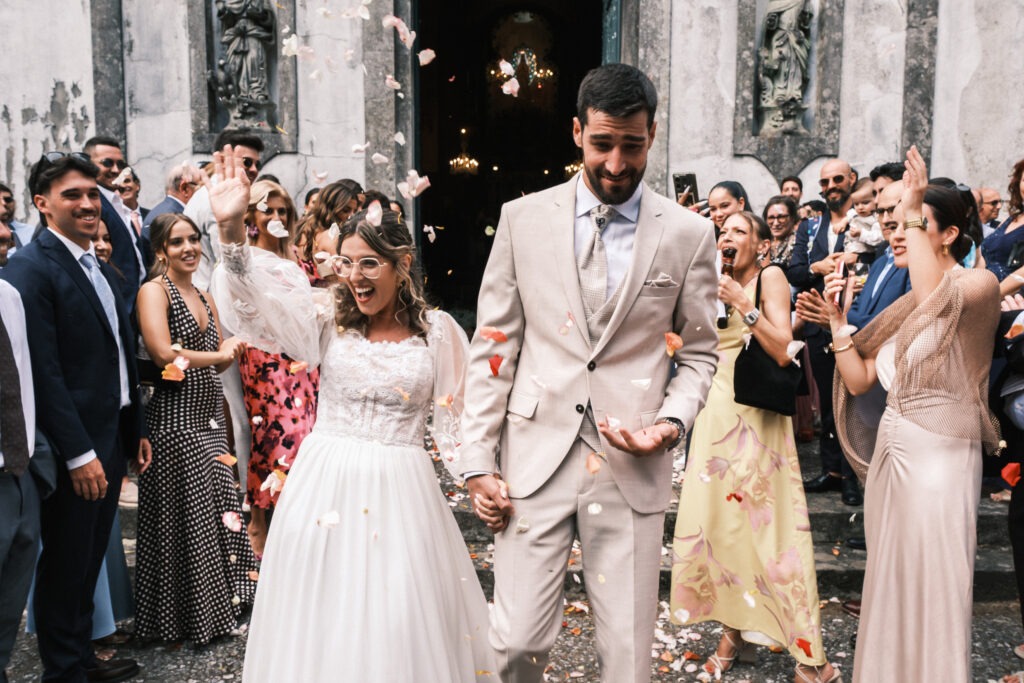Noivos saem da igreja sob uma chuva de pétalas, rodeados por convidados em festa; luz natural, exterior, registo documental em Portugal.
