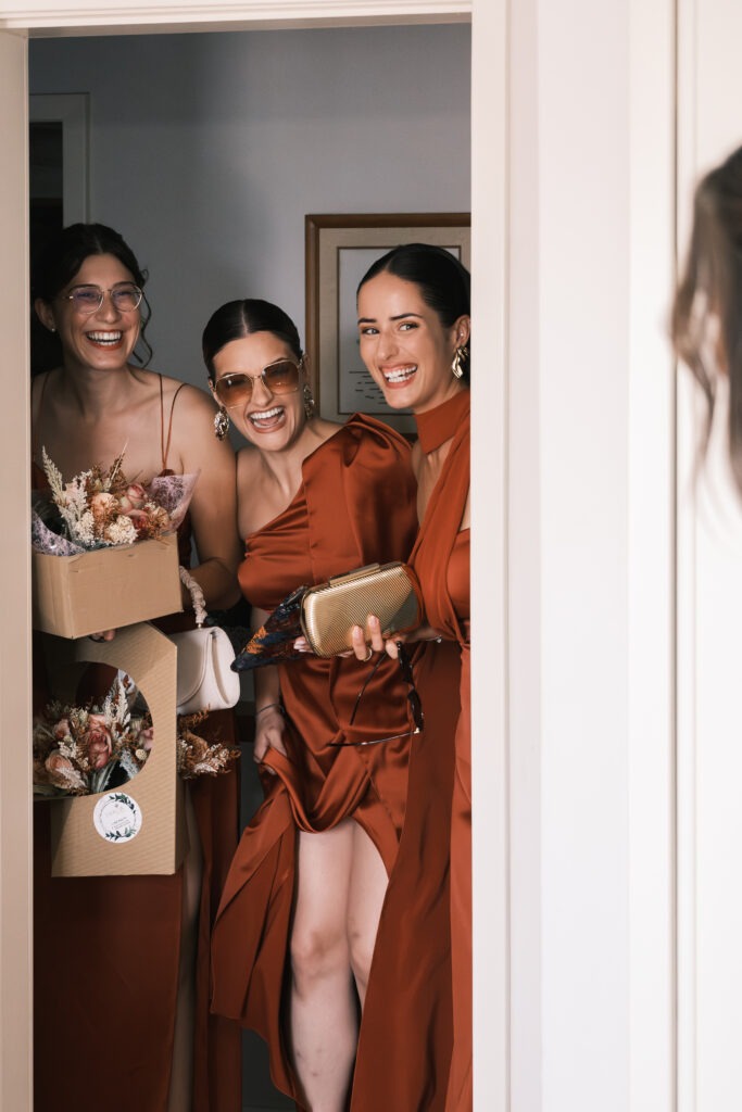 Três madrinhas entram sorridentes com arranjos de flores secas e caixas de bouquets; momento de preparação em interior, luz suave e tom terracota dos vestidos, Santa Maria da Feira.