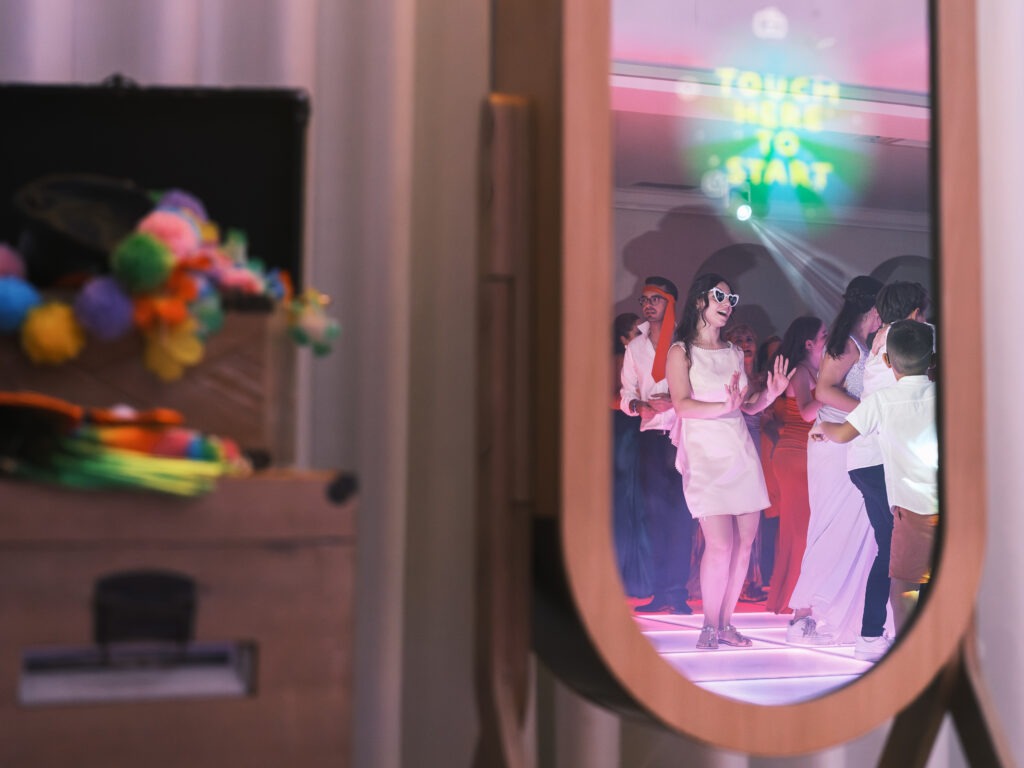 Noiva refletida no espelho do photobooth enquanto dança com os convidados, luzes de festa e ambiente animado em Santa Maria da Feira, registo documental. 
