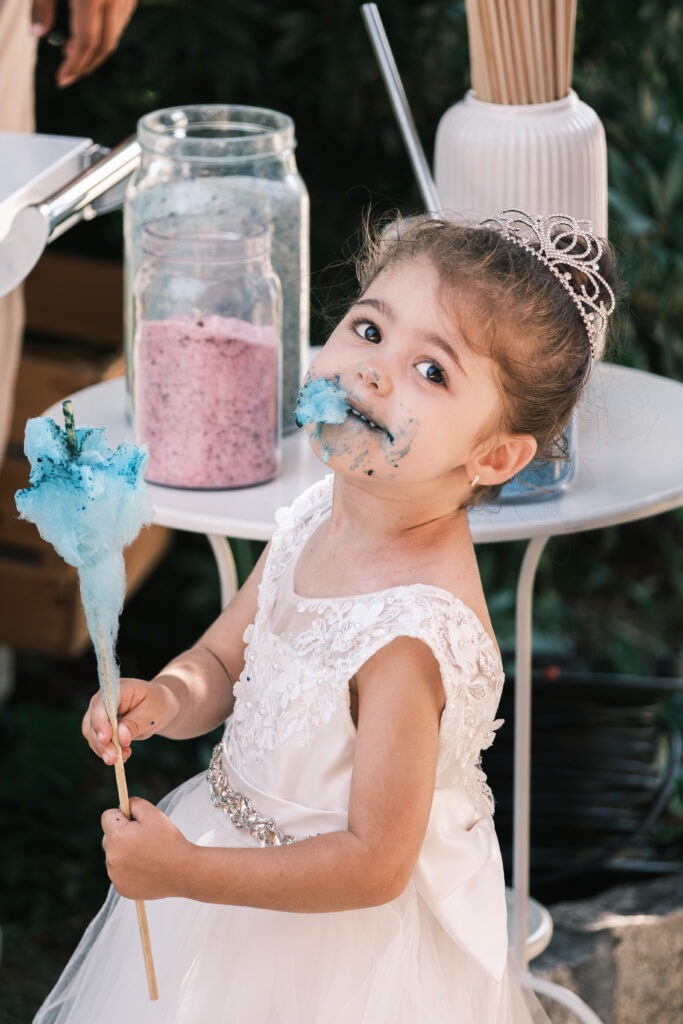 Menina com tiara e vestido branco a saborear algodão doce azul durante o copo-de-água, momento espontâneo ao ar livre em Santa Maria da Feira, registo documental.