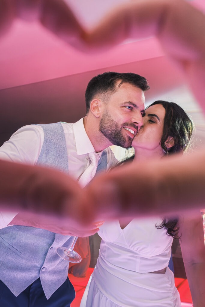 Noiva a beijar o noivo na pista de dança, enquadrados por mãos em forma de coração, luz rosa e ambiente de festa em Aveiro, registo documental.
