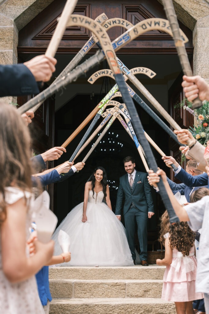 Noivos a sair da igreja sob guarda de honra com sticks de hóquei; amigos e crianças levantam os sticks, ambiente festivo e registo documental em Santa Maria da Feira
