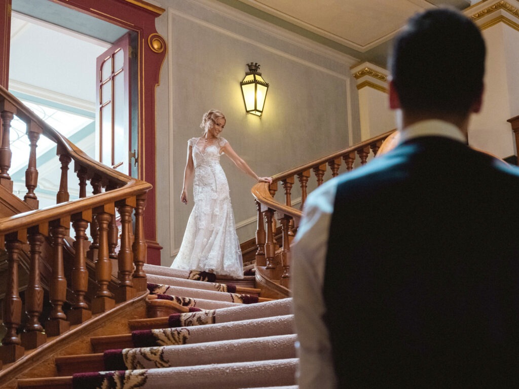 Noiva desce escadaria clássica de madeira com vestido de renda enquanto o noivo a observa em contraluz, momento de first look intimista.