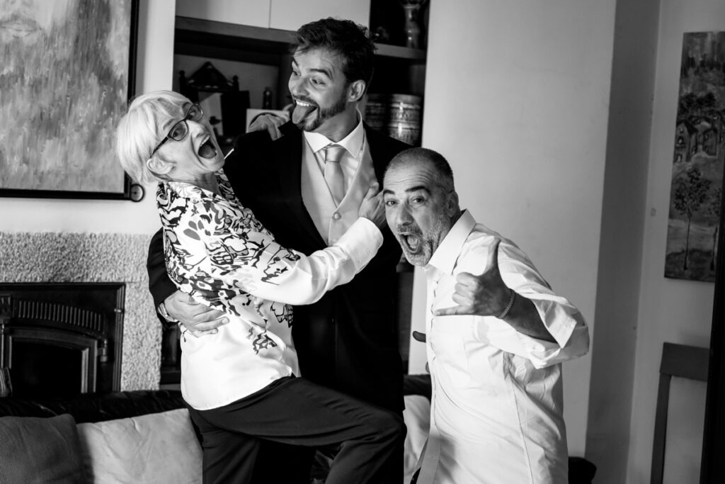 Noivo brinca com os pais com gargalhadas e expressões exageradas durante os preparativos em casa, registo documental em preto e branco.
