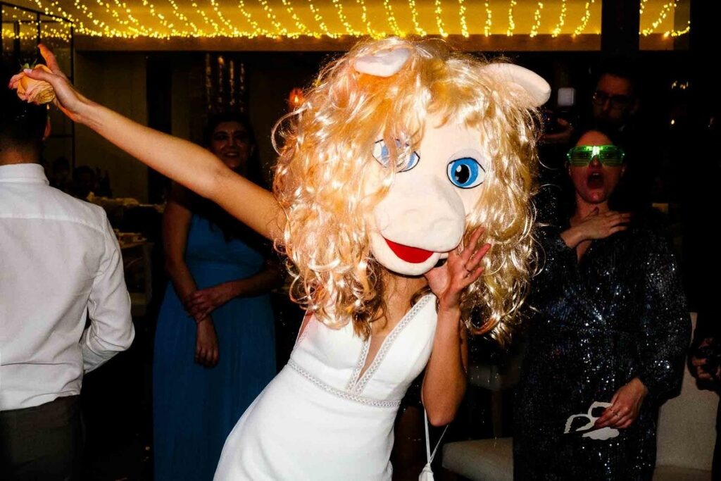Noiva dança com máscara gigante da Miss Piggy durante a festa, luzes amarelas e convidados divertidos no fundo, registo documental nocturno.