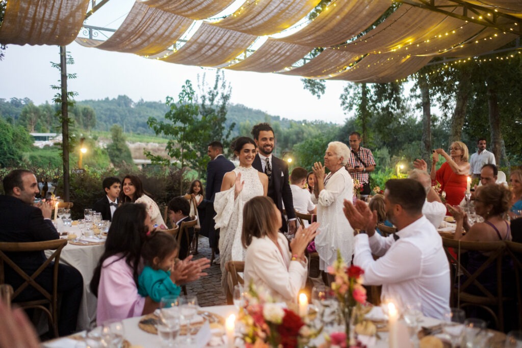 Entrada dos noivos no jantar ao ar livre sob tecidos suspensos e luzes amarelas, convidados aplaudem em pé, registo documental ao fim do dia.