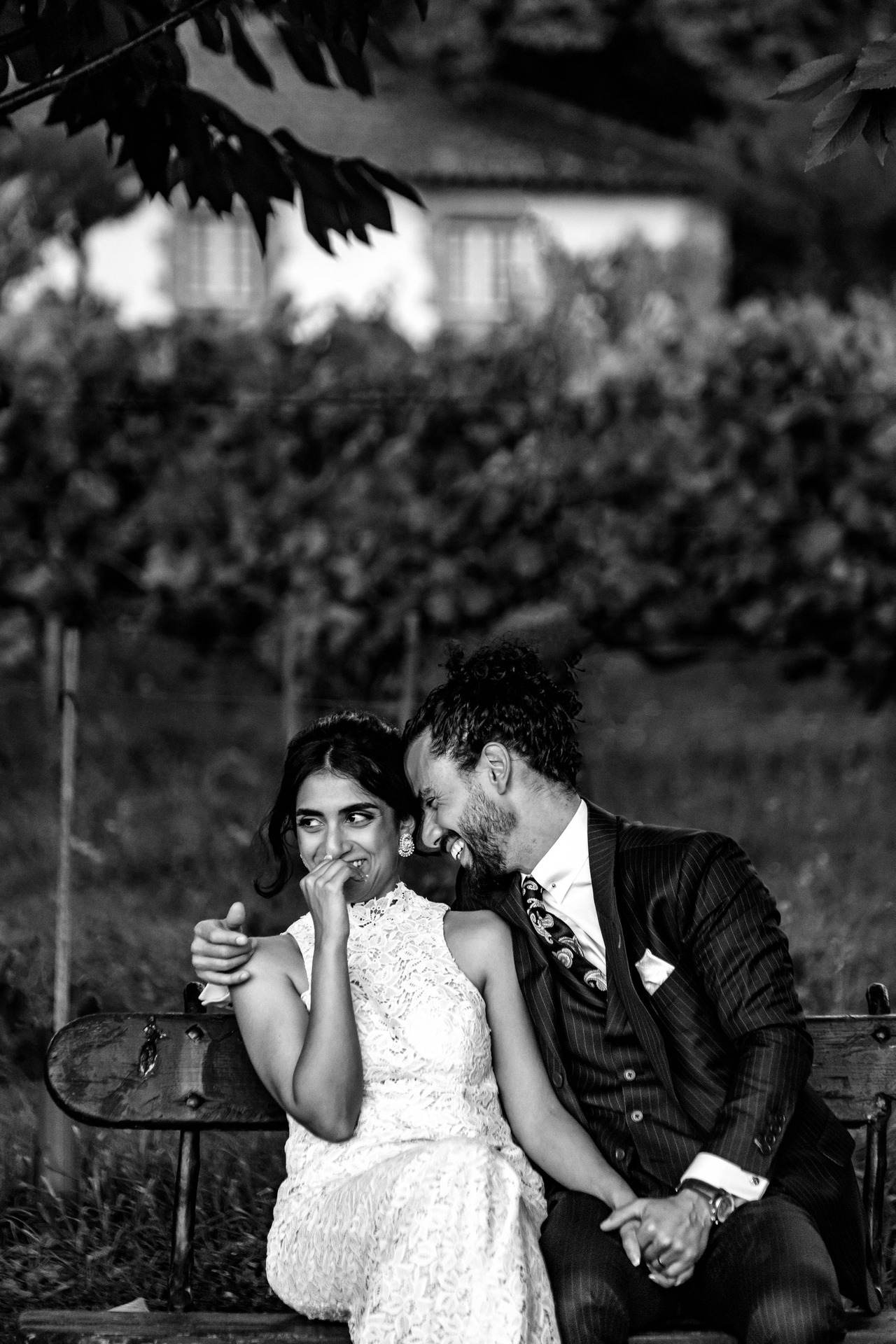 Pequena sessão com os noivos durante o casamento, estando sentados num banco num zona de jardim, noivo encostado à noiva enquanto esta olha para o lado com a mão na boca envergonhada