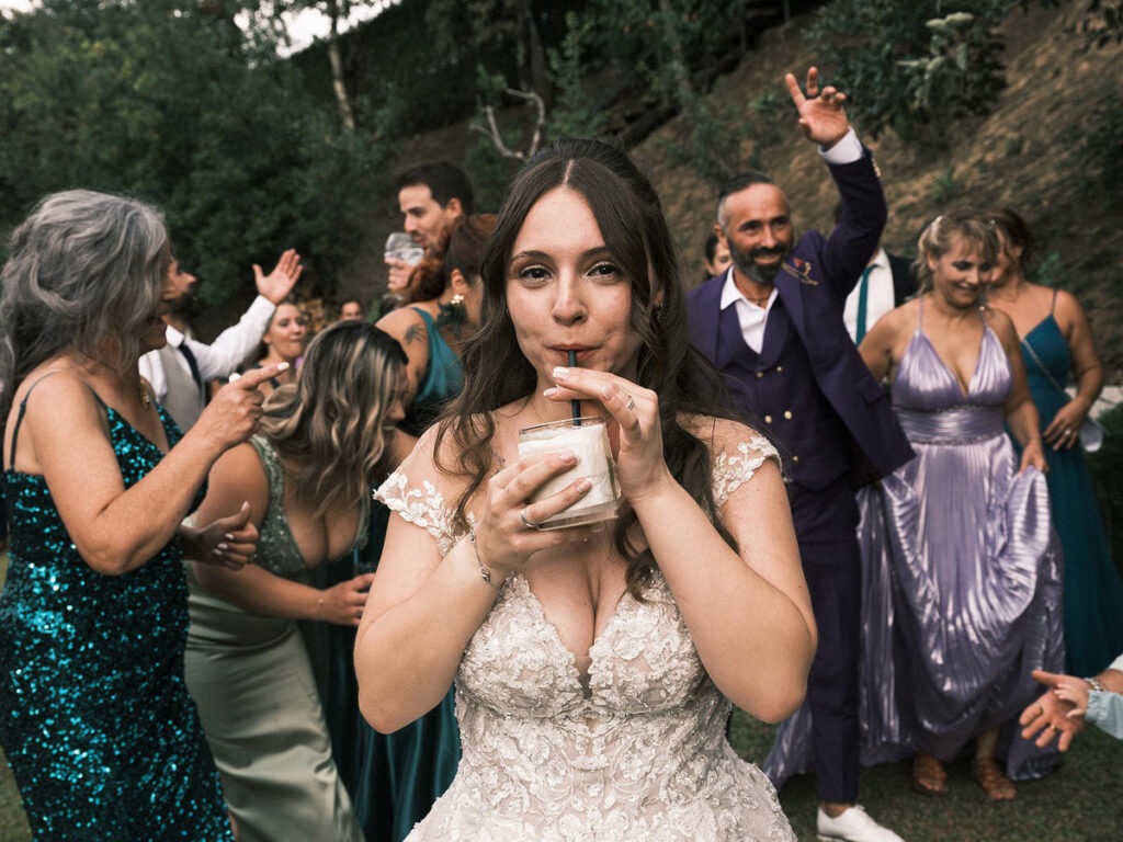 Noiva a beber com palhinha olhando para a câmara enquanto os convidados dançam atrás, momento espontâneo na festa ao ar livre.