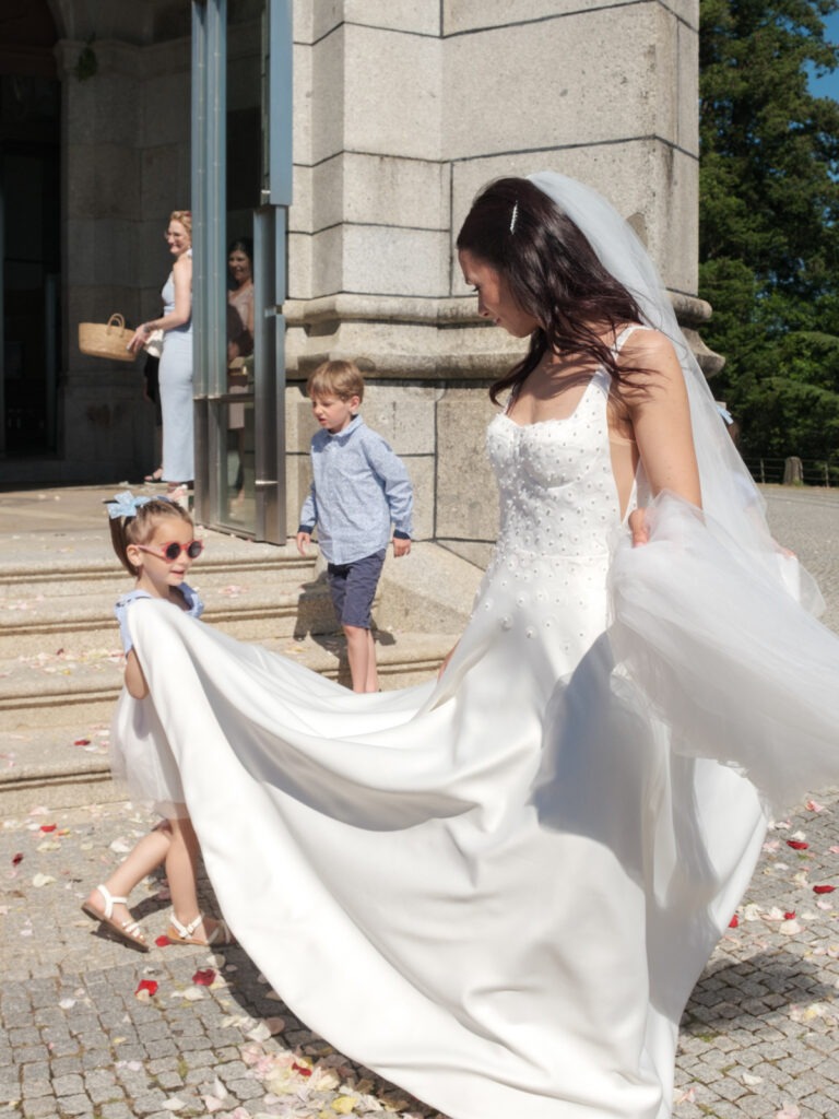 Menina das alianças ajuda a segurar a cauda do vestido da noiva à saída da igreja, pétalas de rosa no chão, registo documental ao sol.