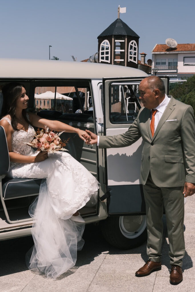 Pai da noiva dá a mão para ela sair da carrinha Volkswagen vintage a caminho da cerimónia, ramo tropical ao colo e sorriso cúmplice.