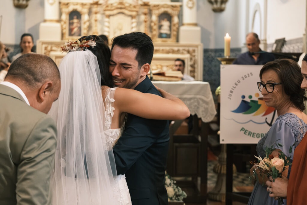 Noivo emocionado com lágrimas abraça a noiva durante a cerimónia em igreja, familiares atentos em redor, registo documental íntimo.
