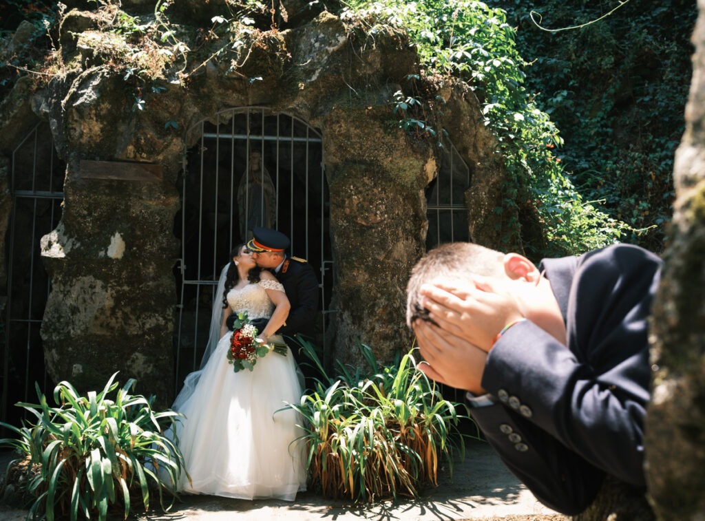 Noivos a beijar-se junto à gruta de Nossa Senhora, noivo de farda militar, rapaz em primeiro plano tapa os olhos com timidez.