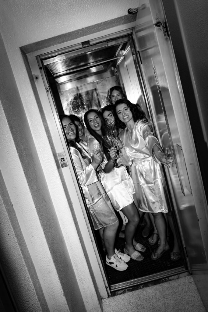 Noiva e amigas de robe de cetim espremidas dentro do elevador a brindar com champanhe, momento divertido dos preparativos em preto e branco.