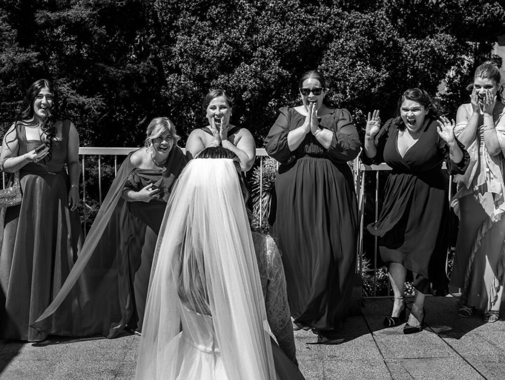 Grupo de amigas reage com surpresa e gargalhadas à noiva num first-look descontraído ao ar livre, registo documental em preto e branco.