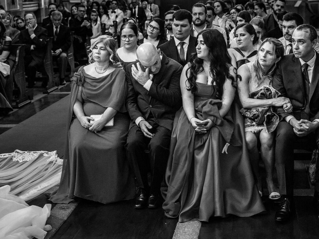 Pai do noivo emocionado cobre o rosto durante a cerimónia enquanto a família observa atenta no banco da igreja, registo documental em preto e branco.