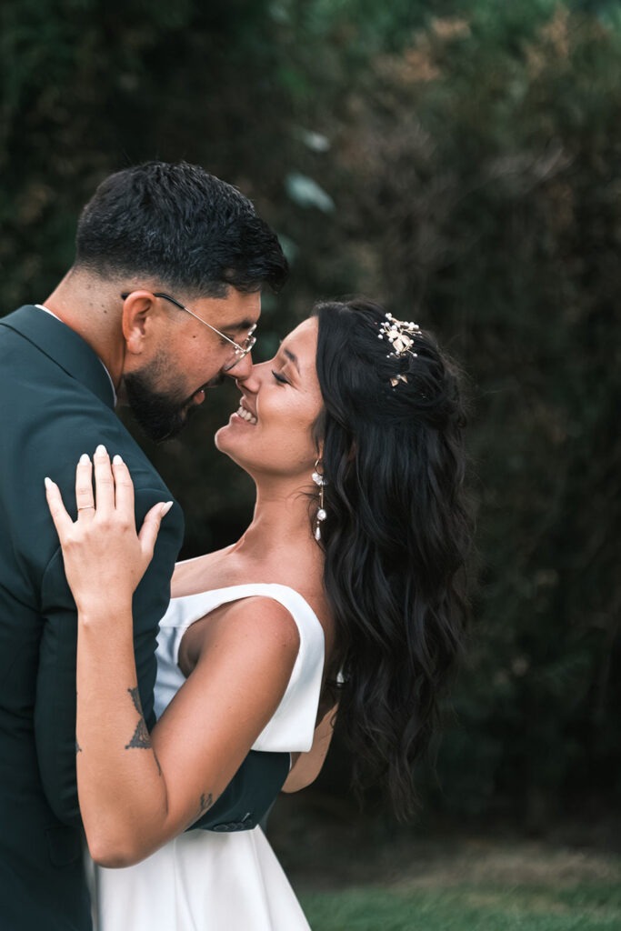 Close-up romântico de noivos testa-com-testa a rir, noiva com cabelo solto e brincos pendentes, fundo de jardim, registo documental.