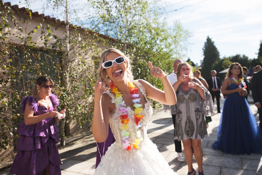 Noiva divertida com óculos de sol e colar havaiano multicolor ri-se durante o cocktail, convidados animados no jardim.