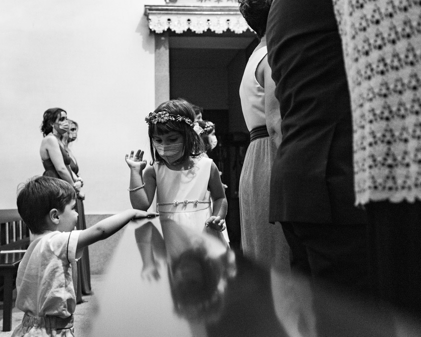 Menina das flores com coroa e máscara cumprimenta menino pequeno na saída da igreja, instante espontâneo em preto e branco.