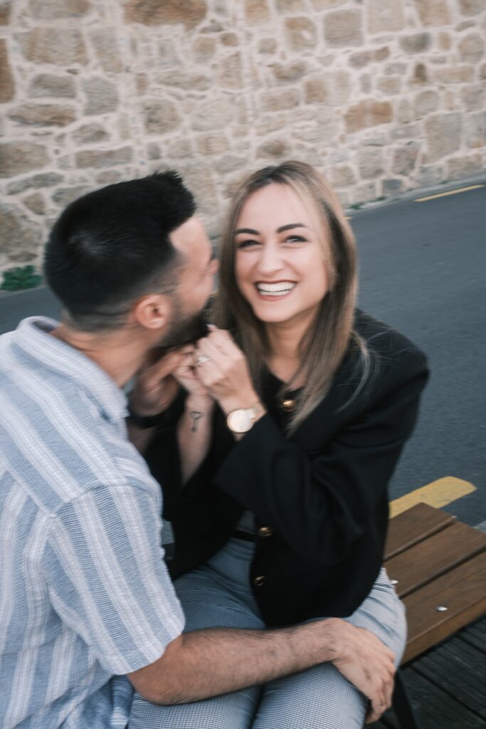 Casal em sessão pré-casamento no Cais de Gaia; noiva puxa a barba do noivo numa brincadeira, riso genuíno, exterior com parede de pedra e banco, luz natural. 
