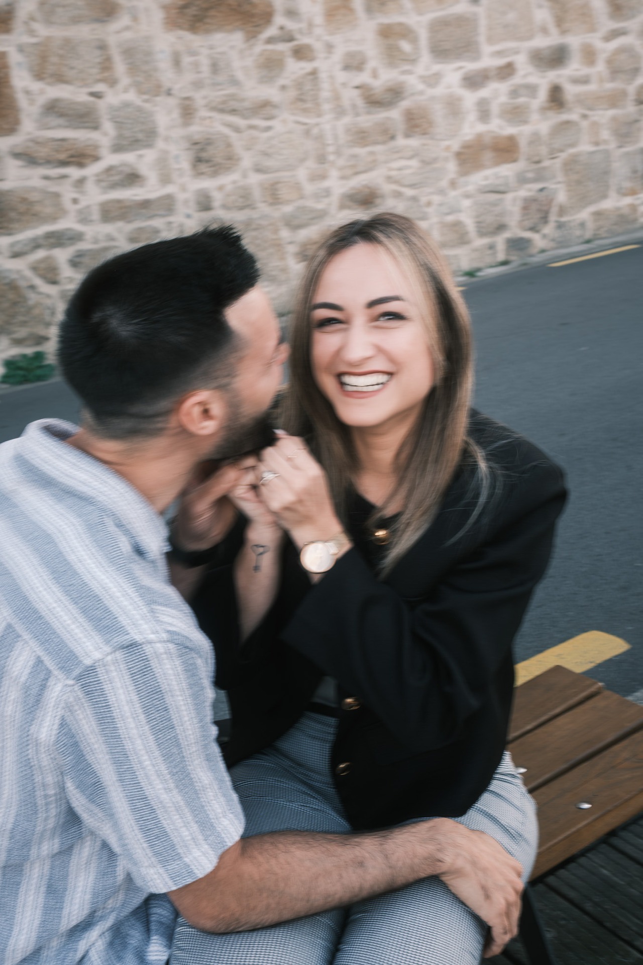 Casal em sessão pré-casamento no Cais de Gaia; noiva puxa a barba do noivo numa brincadeira, riso genuíno, exterior com parede de pedra e banco, luz natural.