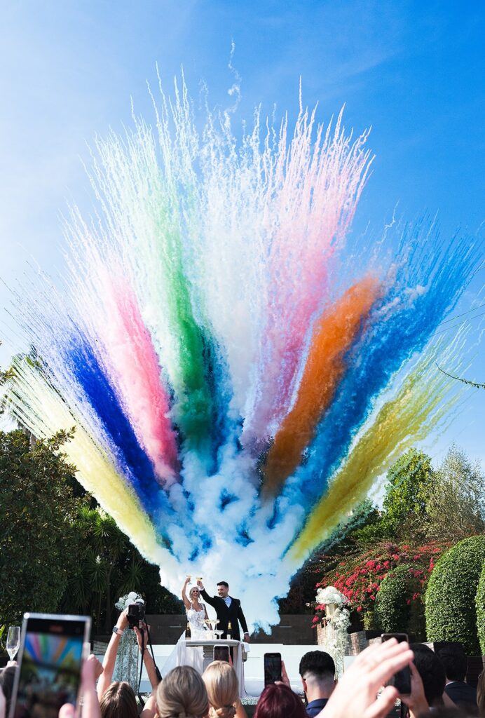 Noivos a brindar e trás deles uma explosão de cores, com os convidados a filmar o momento.